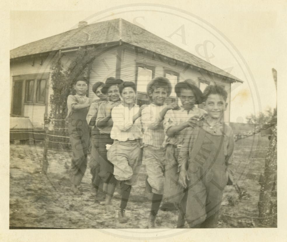 Seven small boys in a line, hands on each other’s shoulders.