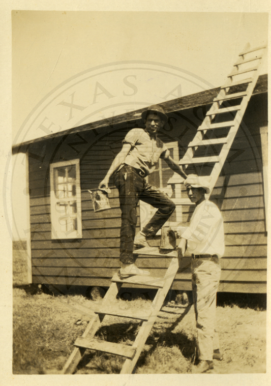 Two men at work on a house, one up a ladder — the school built by its own students.