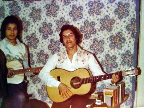Two guitarists in front of a flowered bedspread.