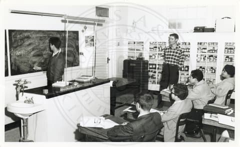 Class. Students sitting in a class and looking at the blackboard. Another student is writing on the blackboard and the teacher is looking at him.