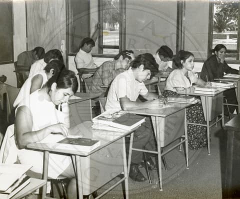 Class. Students sitting in a class with their books opened on their respective desks. A student in the center in the background has raised his hand.