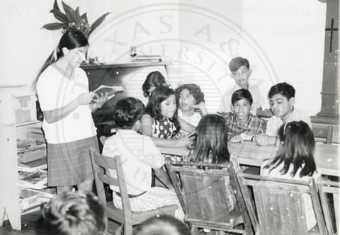 Teacher & Students. A teaching standing & reading from a book. There are small children sitting around a table.