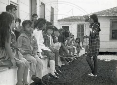 Teacher & Students. Small children sitting outside a house. A teacher is standing in front of them.