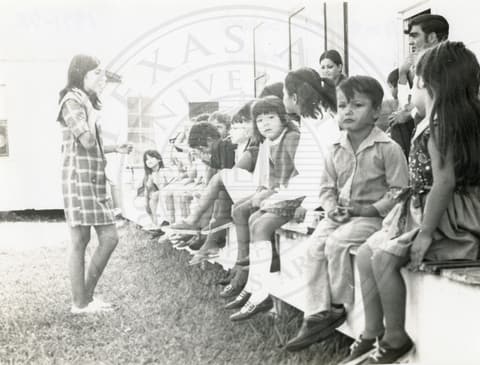 Teacher & Students. Small children sitting outside a house. A teacher is standing in front of them.