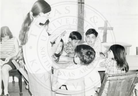 Teacher & Students. Teacher reading from a book in front of small children sitting around a table.