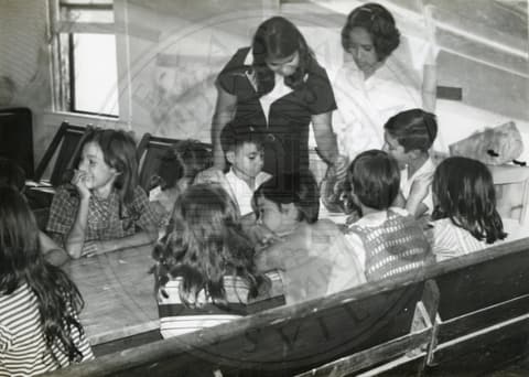 Teacher & Students. Teacher looking at small student's book. Ten students are sitting around a table.